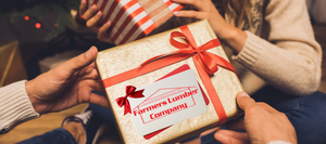 Close-up of two hands exchanging a wrapped gold gift box tied with a red ribbon, featuring a Farmers Lumber Company gift card tucked under the bow. A blurred background shows a cozy holiday setting with a Christmas tree and another gift.